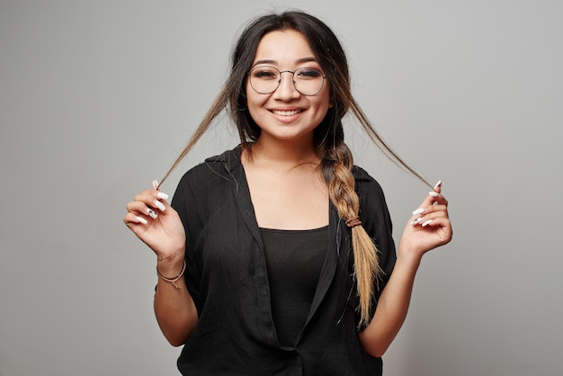 Una studentessa asiatica kazaka intelligente con codino e occhiali tocca i capelli in posa in abiti neri sorridenti su uno sfondo bianco