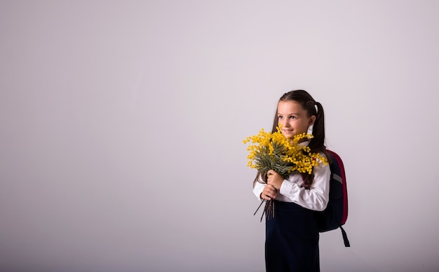 Una studentessa bruna in uniforme e uno zaino tiene un mazzo di mimosa su uno sfondo bianco con un posto per il testo