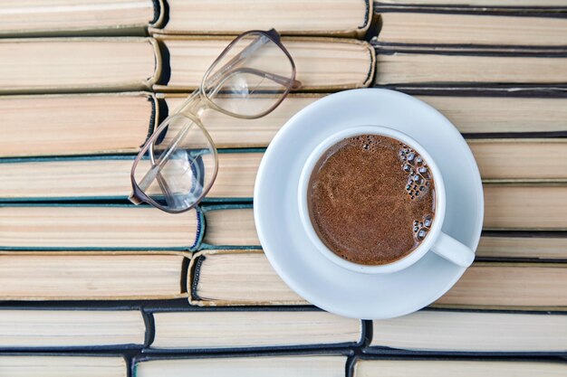 Una tazza bianca di cappuccino profumato al caffè e occhiali da lettura sullo sfondo di vecchi libri usurati impilati a testa in giù