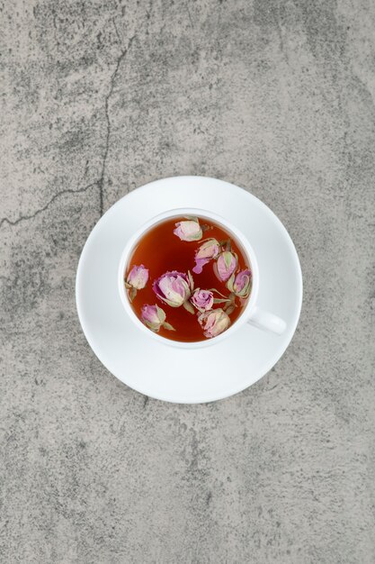 Una tazza bianca di gustoso tè caldo con fiori di rosa appassiti su un tavolo di pietra.