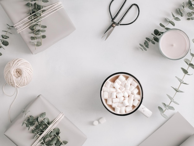 Una tazza con cioccolata calda e marshmallow tra scatole regalo ed eucalipto su un tavolo bianco Il concetto di inverno e regali di confezionamento Vista dall'alto piatta stile minimalista