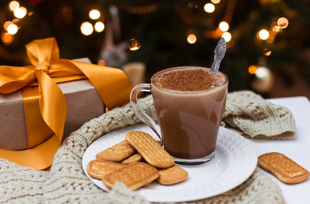 Una tazza di cacao e biscotti su un piatto bianco sul tavolo di un albero di Natale e un regalo