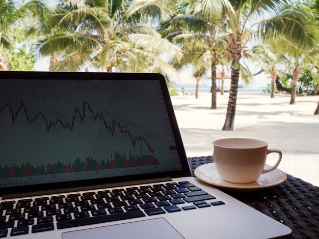 Una tazza di caffè accanto al computer portatile con l'albero di cocco e lo sfondo della spiaggia di sabbia bianca con vista sul mare