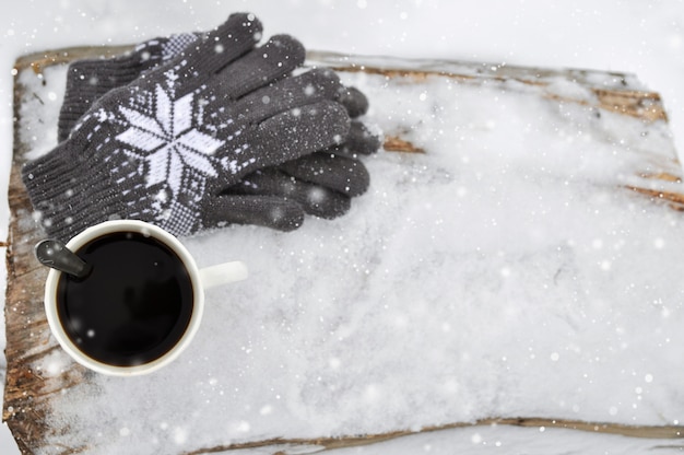 Una tazza di caffè bianca e guanti grigi lavorati a maglia con un modello su un banco di legno nella neve durante precipitazioni nevose.