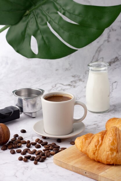 Una tazza di caffè bianco è stata posta accanto a una bottiglia di latte e un cornetto su un tagliere, chicchi di caffè e macinacaffè su un pavimento di marmo.