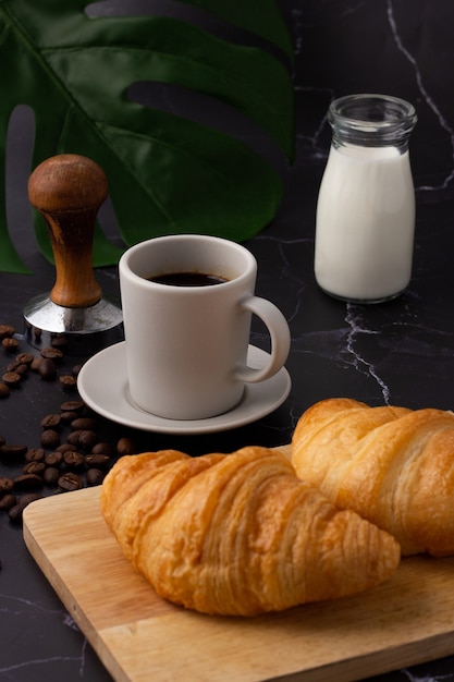 Una tazza di caffè bianco è stata posta accanto a una bottiglia di latte e un cornetto su un tagliere, chicchi di caffè e macinacaffè su un pavimento di marmo.