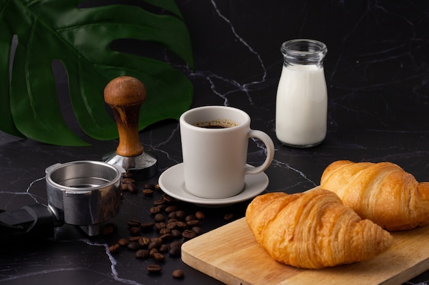 Una tazza di caffè bianco è stata posta accanto a una bottiglia di latte e un cornetto su un tagliere, chicchi di caffè e macinacaffè su un pavimento di marmo.