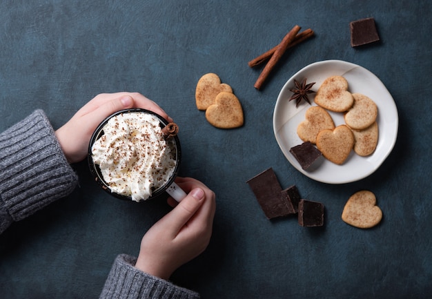 Una tazza di caffè con panna e gocce di cioccolato in mano di donna su un tavolo scuro con biscotti fatti in casa, cioccolato e cannella. Vista dall'alto