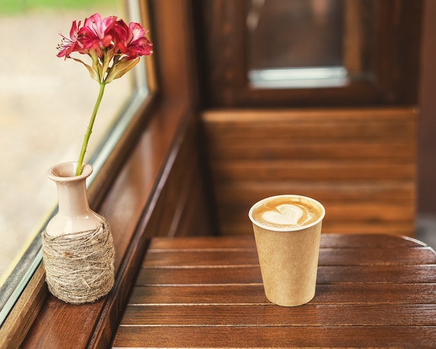 Una tazza di caffè di carta con schiuma a forma di cuore e una brocca con un fiore rosso sul tavolo