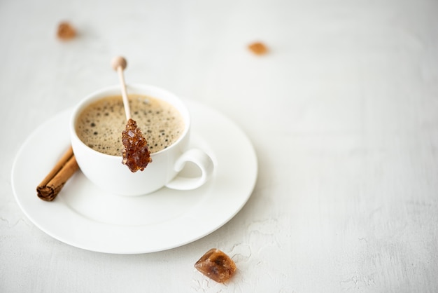 Una tazza di caffè nero con cannella e zucchero caramellato su un bastoncino, su un tavolo bianco