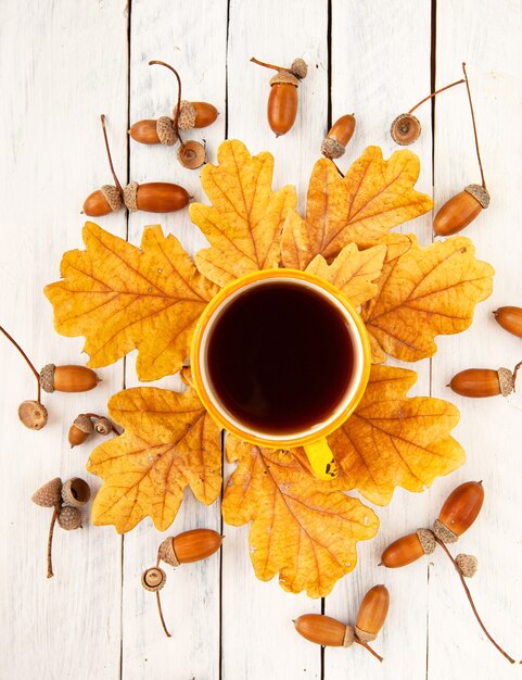 Una tazza di caffè si erge su foglie di quercia gialla autunnale con ghiande su sfondo bianco Vista dall'alto