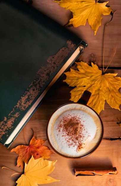 Una tazza di latte con disegno spruzzato accanto a un vecchio libro su un tavolo di legno e foglie d'autunno