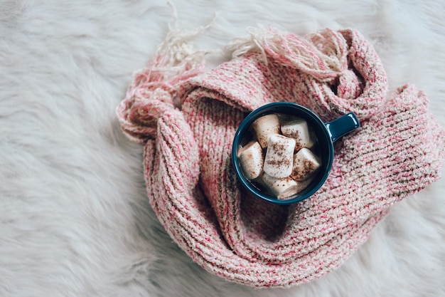 Una tazza di marshmallow al cioccolato caldo circondata da una calda sciarpa lavorata a maglia su un tappeto di pelliccia bianca