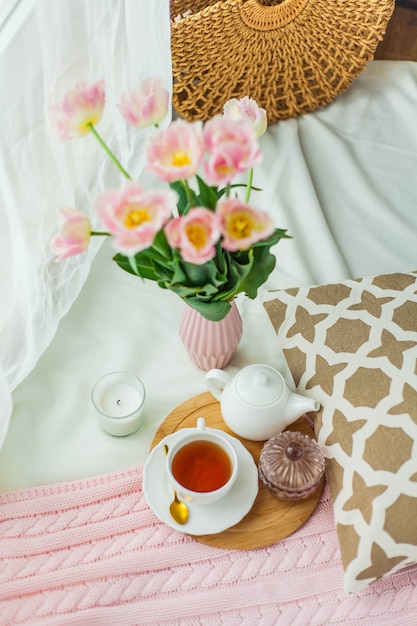 Una tazza di tè caldo, una coperta lavorata a maglia rosa, un mazzo di tulipani in un vaso, una candela sul letto.