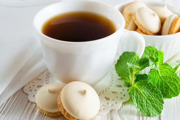 Una tazza di tè e piccoli biscotti con foglie di menta su un tavolo di legno bianco.