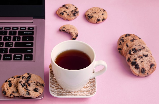 Una tazza di tè è su un piattino vicino a un laptop. Biscotti al cioccolato su uno sfondo rosa.