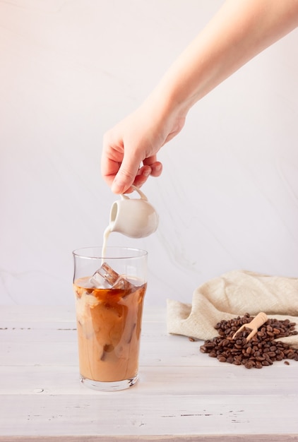 Una tazza di vetro con caffè freddo e ghiaccio, chicchi di caffè su una superficie chiara. Una mano versa la panna e un bicchiere