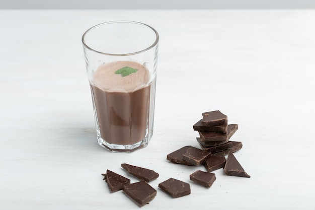 Una tazza di vetro con un frullato proteico al cioccolato si erge sullo sfondo di un pacchetto di proteine su un tavolo bianco. Concetto di nutrizione sportiva.