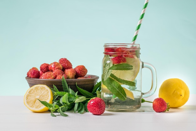 Una tazza di vetro di acqua fredda con menta limone e frutti di bosco su un tavolo di legno bianco