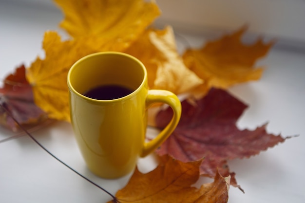 Una tazza gialla con tè e foglie di acero gialle si erge su una finestra bianca in autunno