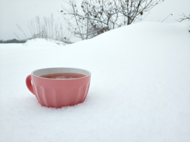 Una tazza rosa con tè caldo al lampone sullo sfondo di un campo invernale innevato. Tè con marmellata riscalda nel freddo inverno.