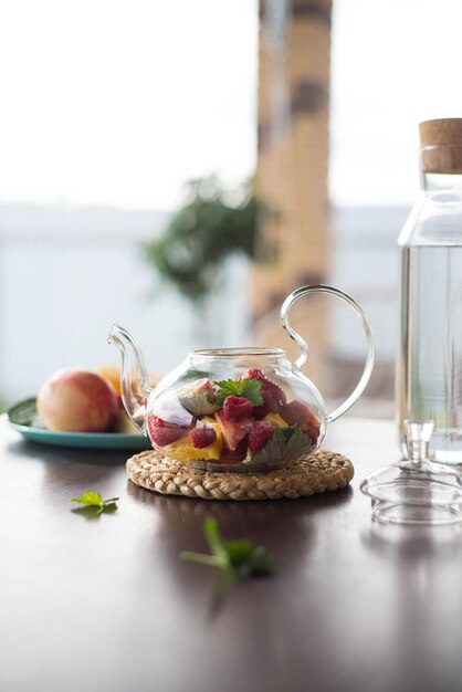 Una teiera di vetro con frutta e bacche di menta lascia una bottiglia d'acqua sul tavolo fuori Tè alla frutta estiva