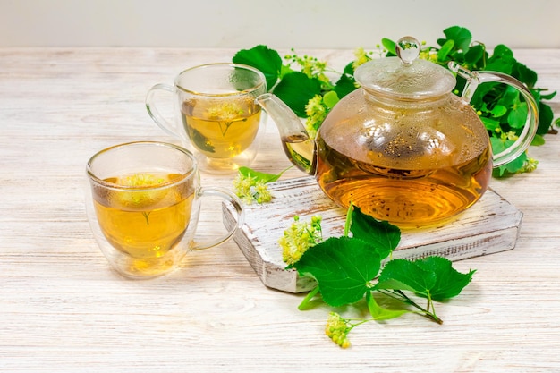 Una teiera di vetro e due tazze di tè con un albero di tiglio su un tavolo di legno sullo sfondo di foglie e fiori di tiglio Tè curativo