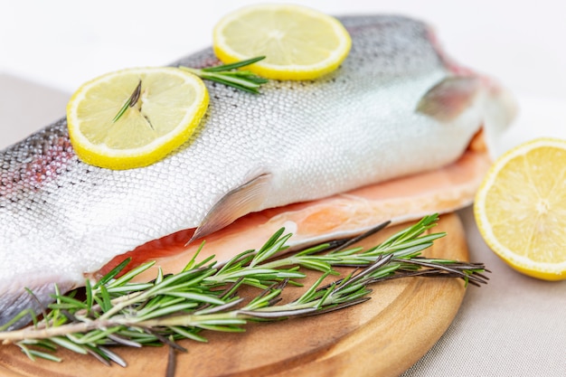 Una trota decapitata cruda affettata su un lato con foglie di limone e rosmarino accanto ad essa. Su un tagliere di legno marrone. Pesce d'acqua dolce.