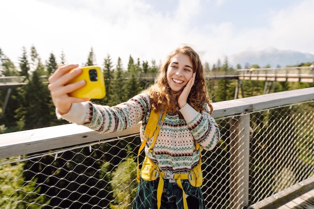 Una turista con uno zaino da escursione si fa un selfie con il telefono su un ponte da escursionismo nell'ambiente forestale