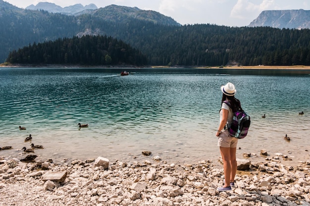 Una turista con uno zaino e un cappello si trova sulla riva di un lago in montagna