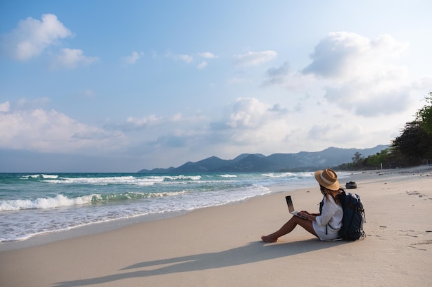 Una viaggiatrice che usa e lavora al computer portatile mentre è seduta sulla spiaggia