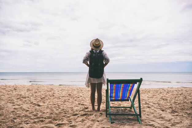 Una viaggiatrice con cappello e zaino in piedi da sola sulla spiaggia in riva al mare