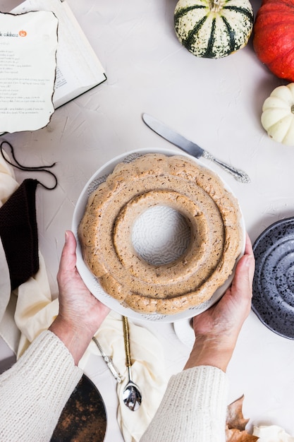 Una vista dall'alto di qualcuno in possesso di una torta di zucca da un tavolo bianco con ingredienti e la ricetta del libro in esso