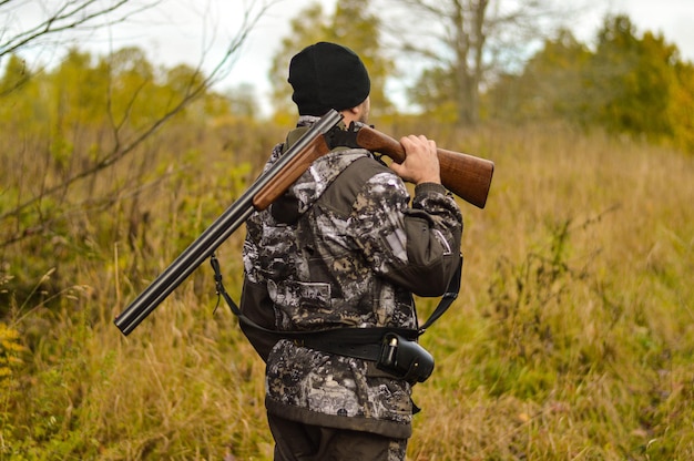 Una vista posteriore di un uomo in uniforme mimetica con una pistola in spalla e una cartuccia che caccia nella foresta autunnale