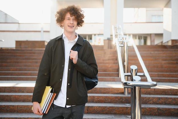 UniversitàSorridente giovane studente uomo con in mano un libro e una borsa su uno sfondo universitario Giovane studente sorridente all'aperto Life styleCityStudent