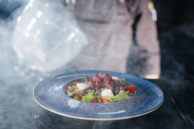 Uno chef professionista serve un'insalata appena preparata di pomodori e verdure di vitello sotto una cappa di vetro con fumo denso