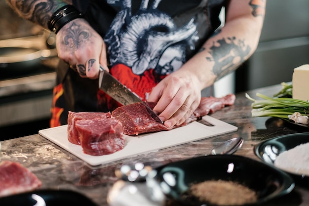 Uno chef tatuato cucina una succosa bistecca a casa Manzo su un tagliere con un coltello e verdure