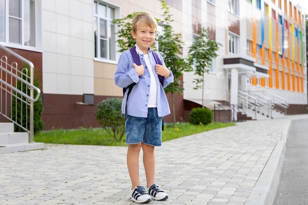 Uno scolaro sta con uno zaino vicino alla scuola, sorride, ride alla scuola elementare