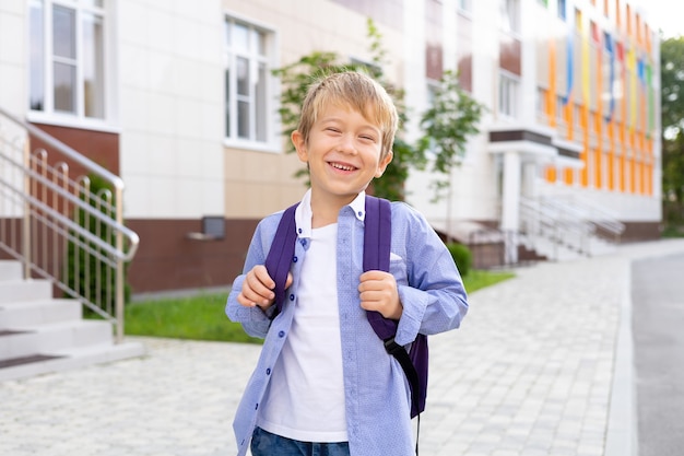 Uno scolaro sta con uno zaino vicino alla scuola, sorride, ride alla scuola elementare