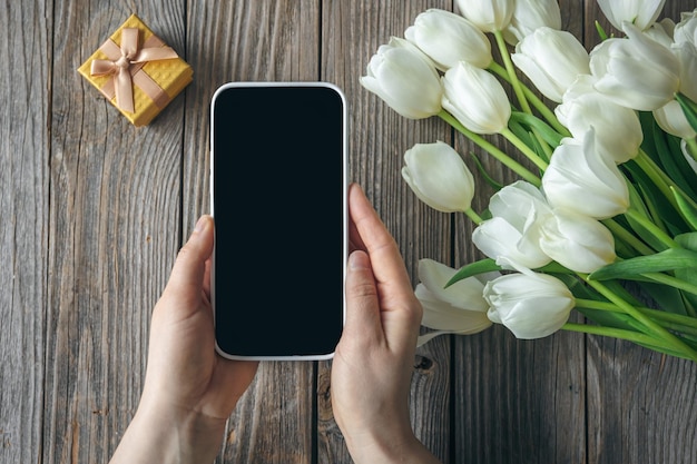 Uno smartphone in mani femminili, una scatola regalo e un bouquet su uno sfondo di legno