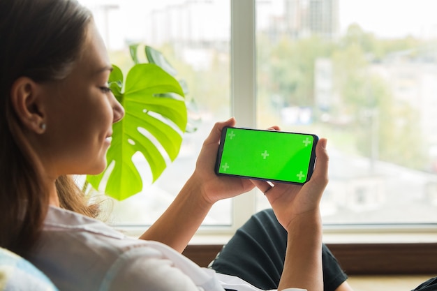 uno smartphone orizzontale con uno schermo chromakey nella mano di una ragazza seduta vicino alla finestra
