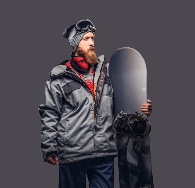 Uno snowboarder rosso brutale con la barba piena in un cappello invernale e occhiali protettivi vestito con un cappotto da snowboard in posa con lo snowboard in uno studio, guardando lontano. Isolato su sfondo grigio.