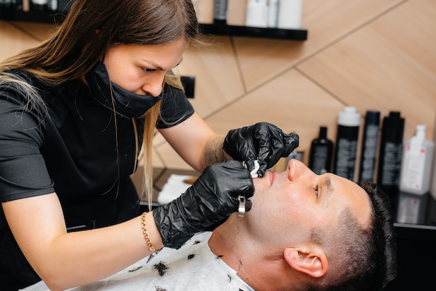 Uno stilista professionista in un barbiere moderno ed elegante rade e taglia i capelli di un giovane uomo. Salone di bellezza, parrucchiere.