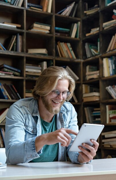 Uno studente felice che usa il tablet guardando lo schermo si siede alla scrivania della biblioteca in verticale