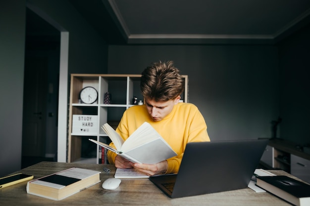 Uno studente in abiti casual luminosi si siede a una scrivania con libri e un laptop in camera da letto e legge attentamente Il giovane studente sta studiando a distanza a casa con libri e Internet School e quarantena