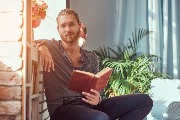 Uno studente rosso bello ed elegante in abiti casual, seduto su una sedia a casa e tiene un libro, guardando una macchina fotografica.