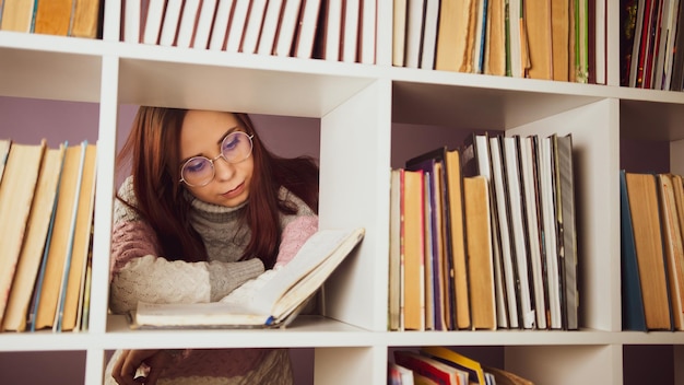 Uno studente sta leggendo un libro in biblioteca dietro gli scaffali Una giovane donna premurosa in piedi in una biblioteca tra libri e leggendo un libro di testo contro un muro viola