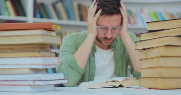 Uno studente stanco si siede alla scrivania piena di libri Giovane uomo che si prepara per un esame nella biblioteca del college Concetto di autoeducazione