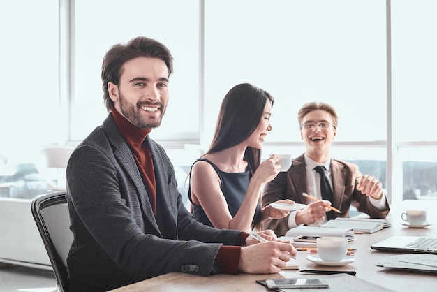 Uomini d'affari e imprenditrice in ufficio che lavorano insieme seduti al tavolo uomo primo piano guardando la telecamera sorridendo felice mentre i colleghi bevono caffè discutendo progetto ridendo