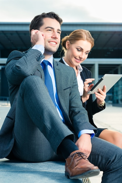 Uomini d'affari o uomo d'affari e donna d'affari che lavorano all'aperto, utilizzando computer pad o tablet e telefono cellulare